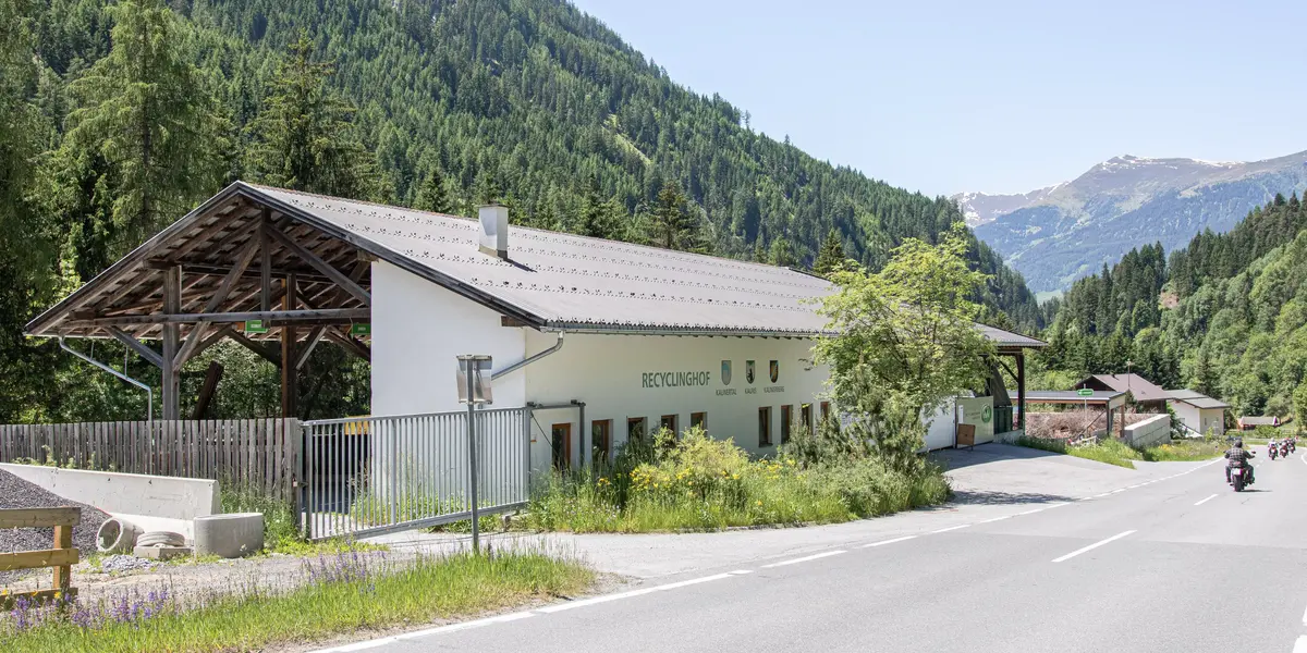 Außenansicht eines hellen Gebäudes mit der Aufschrift "RECYCLINGHOF" entlang einer asphaltierten Straße. Im Hintergrund sind bewaldete Berge unter einem hellen Himmel zu sehen. Ein Motorrad fährt auf der Straße.