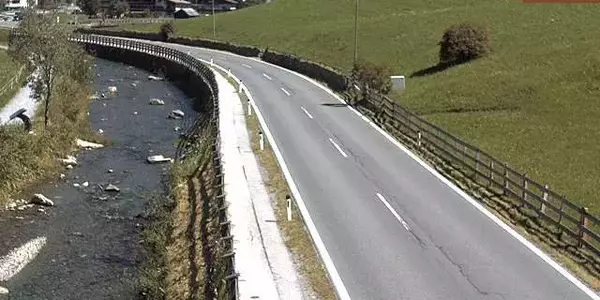 Eine leicht kurvige, zweispurige Asphaltstraße verläuft entlang eines flachen, steinigen Flusses. Links der Straße befindet sich der Fluss mit klarem Wasser und bewachsenem Ufer. Rechts der Straße erstreckt sich ein grüner, leicht abfallender Hang mit ver