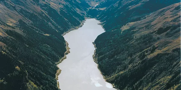 Luftaufnahme eines langen, gewundenen Sees oder Stausees, der von steilen, bewaldeten Bergen umgeben ist. Am unteren Ende ist eine große Staumauer sichtbar. Im Hintergrund ragen schneebedeckte Berggipfel in den Himmel.